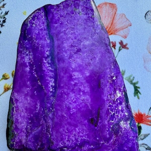 May include: A vibrant, polished purple gemstone with a textured surface. The stone has a natural, irregular shape and displays variations in color and pattern. The gemstone is set against a floral patterned background.