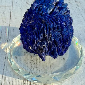 May include: A deep blue azurite mineral specimen with a textured, layered appearance. The mineral sits on a clear, faceted glass dish, reflecting light. The background is a weathered, white wooden surface.