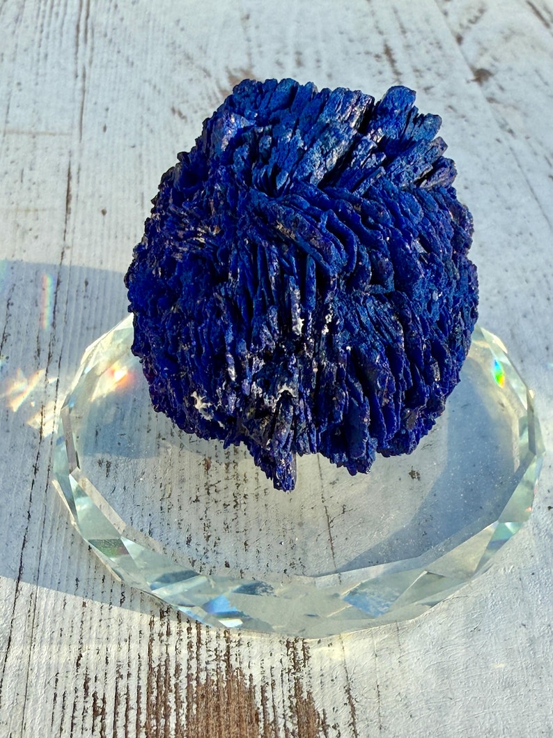 May include: A deep blue azurite mineral specimen with a textured, layered appearance. The mineral sits on a clear, faceted glass dish, reflecting light. The background is a weathered, white wooden surface.
