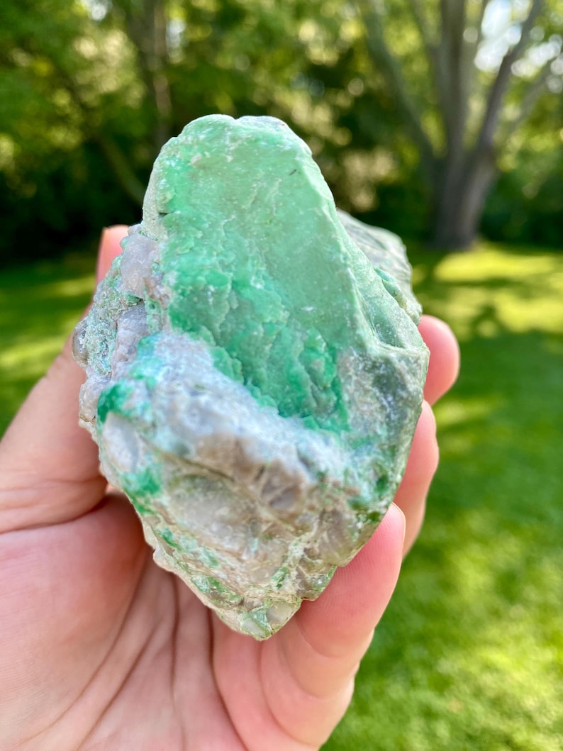 May include: A large, rough, green and gray gemstone with a natural, irregular shape. The stone has a smooth, polished surface and is held in a hand.