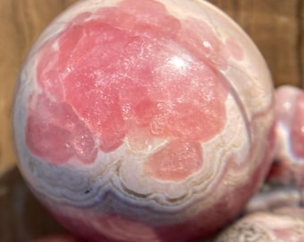 May include: Pink and white banded sphere of polished pink calcite crystal. The sphere has a smooth, polished surface and a natural, organic pattern.