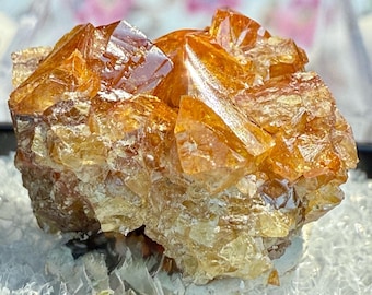 Może przedstawiać: Skupisko żywych, pomarańczowo-żółtych kryształów, prawdopodobnie okaz mineralny. Kryształy mają fasetowaną, geometryczną strukturę, z błyszczącą, odblaskową powierzchnią. Podstawa wydaje się być jaśniejszym, półprzezroczystym materiałem, prawdopodobnie kwarcem.