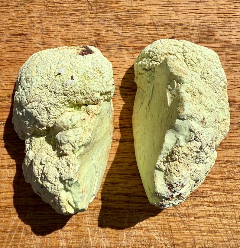 May include: Two irregularly shaped green stones with a rough, textured surface. The stones are light green in color and have a slightly yellowish hue. The stones are placed side-by-side on a wooden surface.