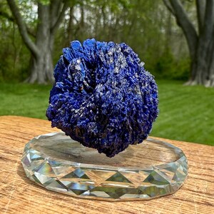 May include: A vibrant blue azurite mineral specimen with a crystalline structure, displayed on a clear, faceted crystal base. The azurite has a textured surface and is set against a blurred green background.