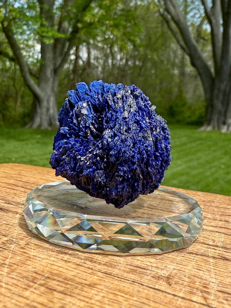 May include: A vibrant blue azurite mineral specimen with a crystalline structure, displayed on a clear, faceted crystal base. The azurite has a textured surface and is set against a blurred green background.
