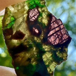 May include: A translucent, irregularly shaped stone with a mix of colors. The stone features areas of deep green, reddish-purple, and a yellowish-brown hue. The stone's surface has a textured appearance, with visible patterns and variations in color.