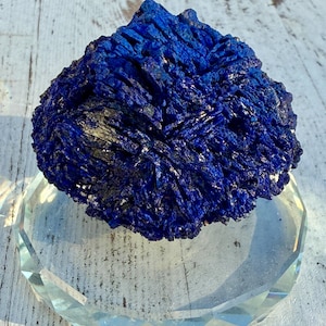 May include: A vibrant blue azurite mineral specimen rests on a clear, faceted glass display stand. The azurite has a crystalline structure with a deep, rich color. The stand is round with a geometric design, set against a white wooden surface.