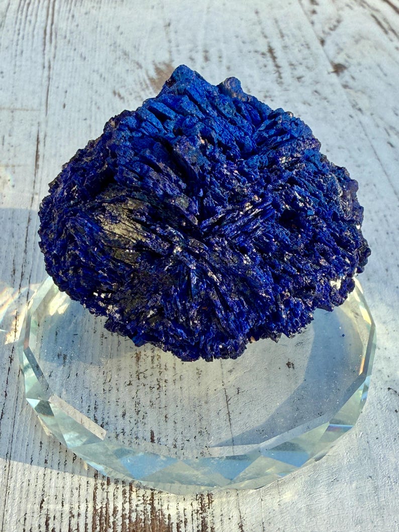 May include: A vibrant blue azurite mineral specimen rests on a clear, faceted glass display stand. The azurite has a crystalline structure with a deep, rich color. The stand is round with a geometric design, set against a white wooden surface.