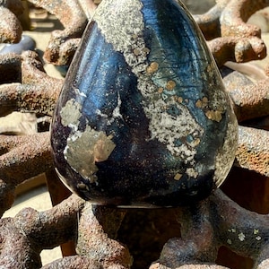May include: A dark blue and silver gemstone with gold flecks, shaped like an egg, resting on a rusty metal surface.
