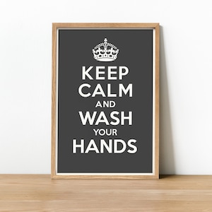 May include: A framed print with a dark gray background and white text that reads "KEEP CALM AND WASH YOUR HANDS." A white crown is at the top. The frame is light brown, and the print is on a wooden surface.