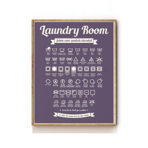 May include: A purple poster with a white border and the text "Laundry Room" in a white vintage font. The poster features a chart of laundry care symbols with their meanings, including washing, drying, ironing, and bleaching instructions. The text "fabric care symbols decoded" is also included in white.