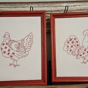 May include: Two framed embroidery pieces with red frames. The left piece features a red embroidered chicken with a white background. The right piece features a red embroidered rooster with a white background.