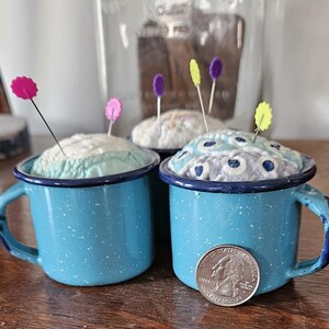 May include: Three blue enamel mugs with handles, each filled with a fabric pincushion topped with colorful pins. The mugs are on a wooden surface.