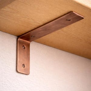 Peut inclure: Un support d'étagère en métal de couleur cuivre est fixé à une étagère en bois de couleur claire. Le support est en forme de L et fixé avec des vis. Le support est conçu pour soutenir une étagère et est monté sur un mur blanc.