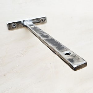 Op de afbeelding: Een zilveren metalen plankbeugel met een T-vormige basis en een lange, rechte arm. De beugel is gemonteerd aan een witte muur.