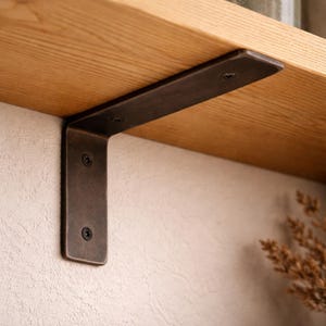 May include: A dark bronze-colored metal shelf bracket is mounted under a light-colored wooden shelf. The bracket is L-shaped and secured with screws. The shelf is made of light-colored wood.