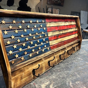 May include: Wooden key holder with an American flag design. The flag is painted in red, white, and blue with a distressed look. The key holder has five hooks.