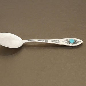 May include: A silver spoon with an oval bowl and a decorative handle. The handle features etched designs and a turquoise-colored stone at the end. The spoon is displayed on a brown surface.