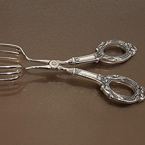 May include: Silver-toned serving tongs with ornate handles and a fork-like end. The tongs are designed with a scissor-like mechanism and are likely used for serving food. The handles have a decorative floral design.