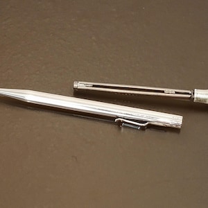 May include: A silver mechanical pencil with a clip and a separate, smaller component. The pencil is lying on a dark brown surface. The pencil's tip is pointed, and the design is sleek and modern.