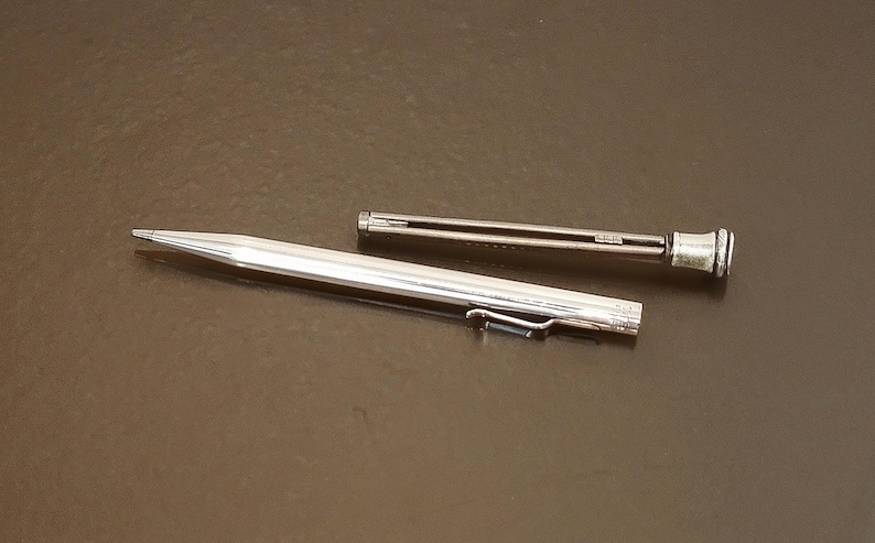 May include: A silver mechanical pencil with a clip and a separate, smaller component. The pencil is lying on a dark brown surface. The pencil's tip is pointed, and the design is sleek and modern.