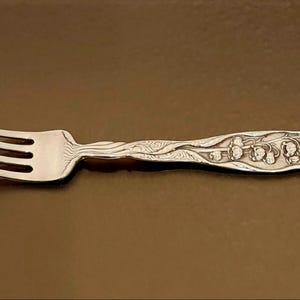 May include: A silver fork with an ornate design on the handle. The fork has four tines and is in focus against a brown background.