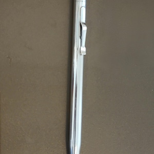 May include: A shiny, silver mechanical pencil with a clip and a textured top. The pencil is slender and tapers to a fine point. The background is a solid, neutral brown.