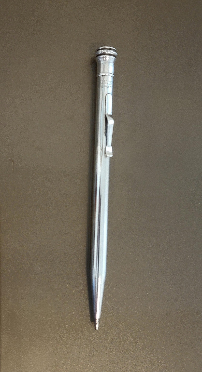May include: A shiny, silver mechanical pencil with a clip and a textured top. The pencil is slender and tapers to a fine point. The background is a solid, neutral brown.