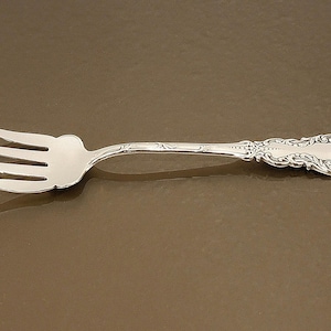 May include: A silver-toned metal fork featuring four tines and an ornate, embossed handle with intricate scrollwork and decorative detailing. The utensil has a polished finish and a curved neck leading to a wide, patterned base on the handle.