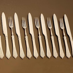 May include: A collection of silver-colored forks and knives arranged on a brown surface. The silverware features ornate floral designs on the handles. The knives have a smooth, curved blade. The forks have three tines.