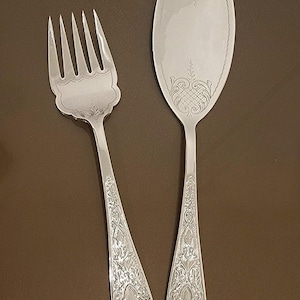 May include: Silver serving fork and spoon set. The fork has four tines, and the spoon is oval-shaped. Both pieces have ornate, engraved floral and geometric patterns on the handles and serving surfaces.