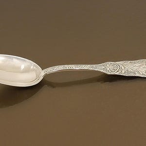May include: A silver spoon with an oval bowl and a decorative handle. The handle features an ornate floral design. The spoon is lying on a brown surface, reflecting the light.