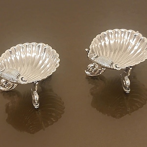 May include: Two silver-colored, shell-shaped dishes with scalloped edges. Each dish is supported by three ornate legs and features a decorative fish-like accent. The dishes are displayed on a reflective, brown surface.