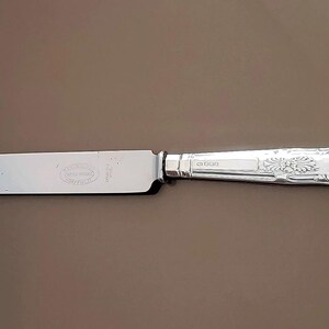 May include: A silver butter knife with an ornate handle and a plain blade. The handle has a floral design and a shell-shaped end.
