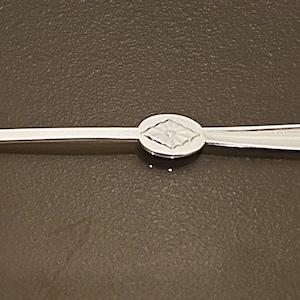 May include: A silver-toned metal seafood fork featuring a slender, two-pronged tip on one end and a rounded, spoon-like handle on the other. The center of the utensil is adorned with a circular medallion embossed with a diamond-shaped pattern.