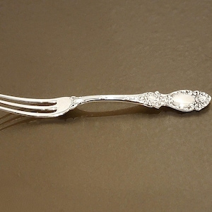 May include: A silver fork with three tines and ornate detailing on the handle. The handle features a floral design and a small, oval-shaped embellishment. The fork is set against a brown background.