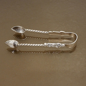 May include: Silver sugar tongs with shell-shaped spoon ends and a twisted handle design. The tongs have a decorative floral motif on the handle. The tongs are displayed on a reflective surface.