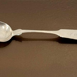 May include: A silver teaspoon with a rounded bowl and a long, slender handle. The handle has a decorative design and is engraved with the letter "J".