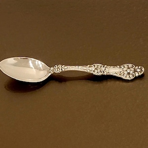 May include: A silver teaspoon with an ornate handle. The spoon has a rounded bowl and a decorative design on the handle.