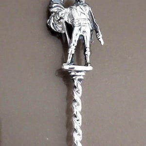 May include: A silver spoon with a twisted handle and a figurine of a person holding a flag on top.