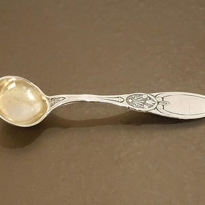 May include: A silver spoon with an oval bowl and a decorative handle. The handle features an ornate design and an oval area for engraving. The spoon is likely vintage or antique.
