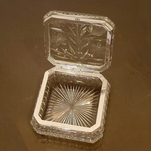 May include: A clear, rectangular crystal box with a hinged lid, featuring a silver-toned metal frame. The lid is open, revealing etched floral designs and a starburst pattern on the base. The box is likely for jewelry or small keepsakes.