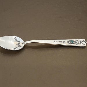 May include: A silver spoon with intricate black and turquoise designs. The spoon features an eye motif and other geometric patterns along the handle. The spoon's bowl is smooth and reflective, and the handle is detailed.