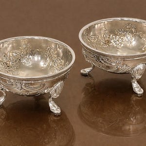 May include: Two antique silver bowls with ornate floral designs and three decorative feet. The bowls have a reflective surface and are set against a brown background. The bowls are likely for serving or display.