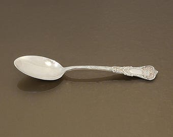 May include: A silver spoon with an ornate handle and oval bowl. The spoon is lying on a brown surface. The handle features a detailed floral design and an oval cartouche.