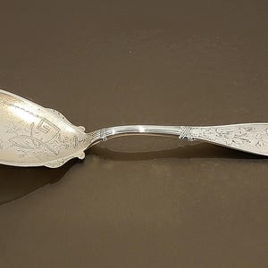 May include: A silver spoon with an ornate floral design on the bowl and handle. The spoon has a curved handle and a wide, oval bowl. The spoon is likely made of sterling silver or a similar metal. The spoon is a vintage or antique item.
