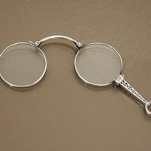 May include: Silver-toned, round eyeglasses with a handle. The glasses have two circular lenses connected by a curved bridge. The handle features an ornate, openwork design and a small ring at the end.