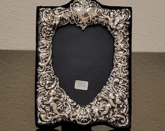 May include: A rectangular silver-toned picture frame featuring an ornate relief design of cherubs, scrolling foliage, and a central face at the bottom. The frame has a dark interior border and a small white sticker reading MADE IN ENGLAND.