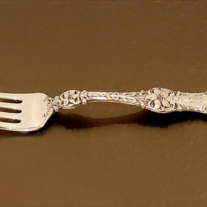 May include: A silver fork with a decorative floral pattern on the handle. The fork has four tines and is set against a brown background. The handle features intricate detailing.