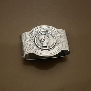 May include: A silver napkin ring with an embossed design. The central oval features a profile of a woman in a laurel wreath. The ring has decorative patterns on the sides and top, with a polished finish.